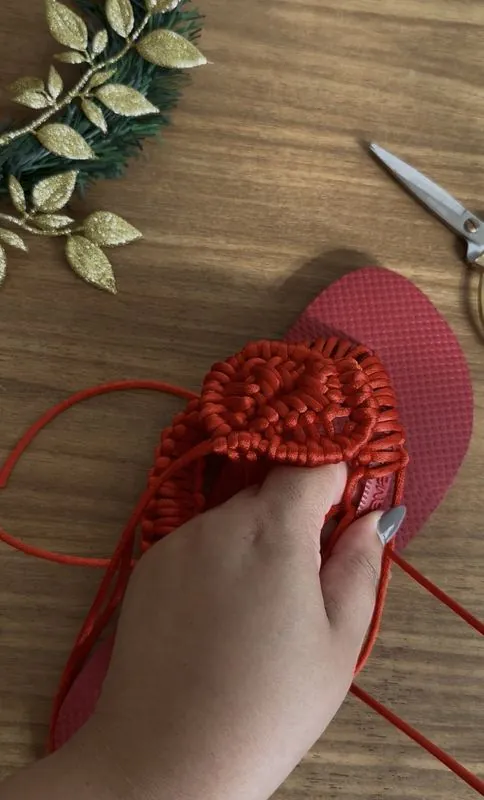 Mãos da Lari tecendo uma sandália de macramê vermelha